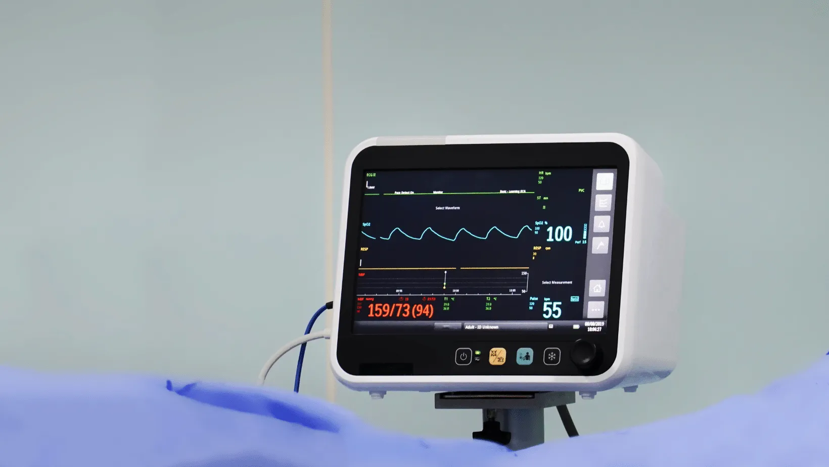 A patient monitor displaying vital signs including ECG, SpO2, and blood pressure with numerical values, set against a pale background.