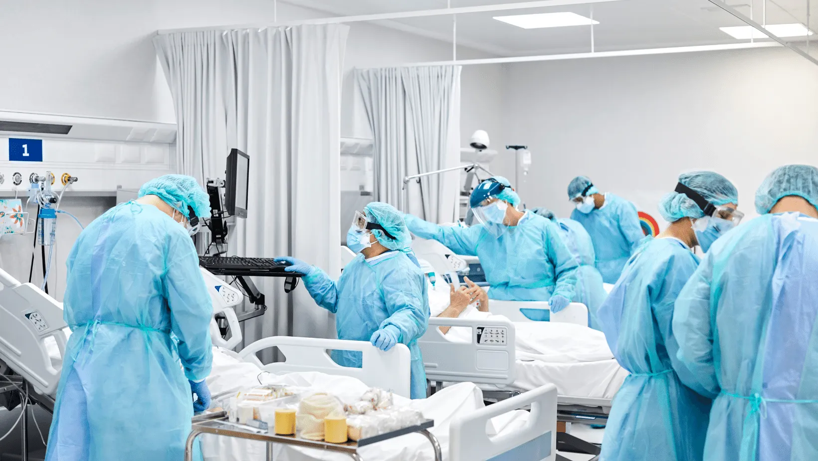 Medical staff in scrubs and protective gear attending to patients and equipment in a busy hospital intensive care unit.