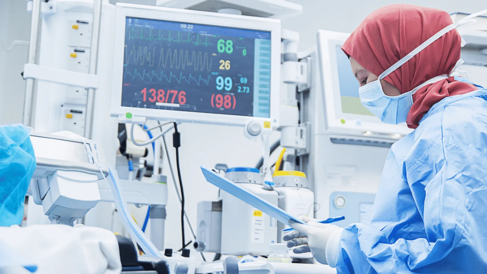 Healthcare professional in blue scrubs and hijab focusing on a patient chart in an ICU, with a vital signs monitor displaying ECG, oxygen saturation, and blood pressure in the background.